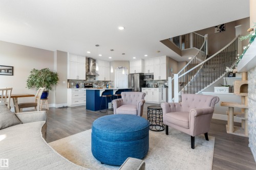 Spacious open-concept living area featuring wood-finish flooring, a contemporary kitchen with white cabinetry and a blue island, and a prominent staircase with dark metal balusters - 3334 Parker Loop Sw, Edmonton, AB 