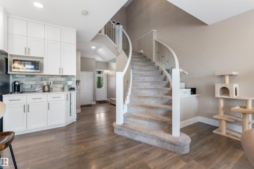 Curved carpeted staircase with white balusters, wood-finish flooring, white kitchen cabinetry, and stone-tiled backsplash - 3334 Parker Loop Sw, Edmonton, AB 